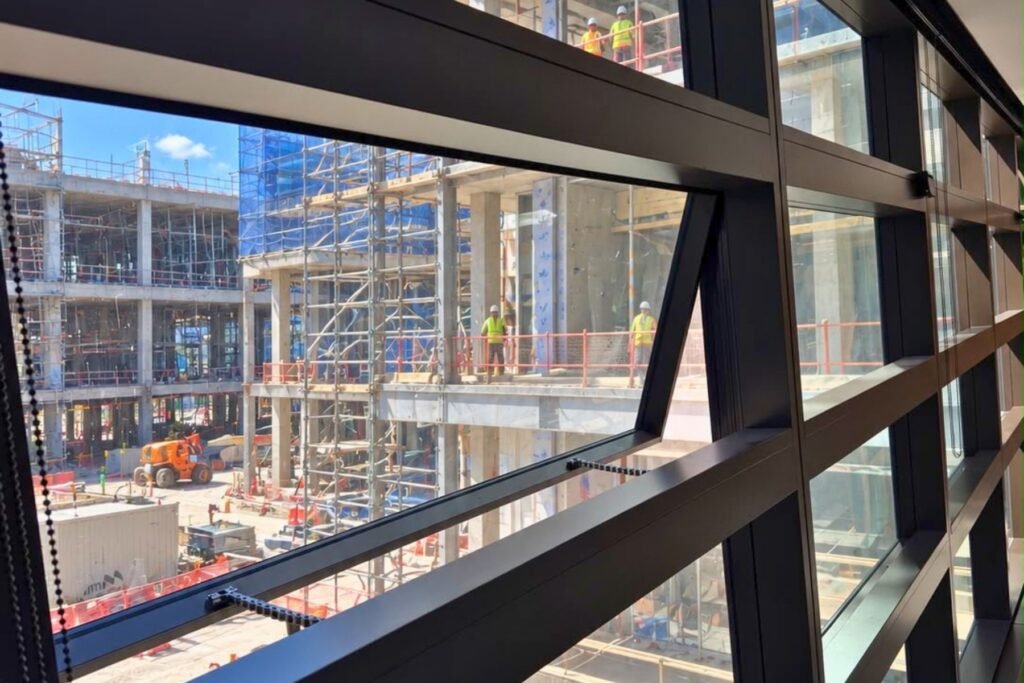 Open top-hung window with two chain actuators, overlooking an active construction site